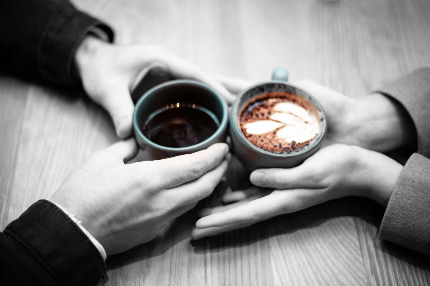Two people hold their coffee cups together across a wooden table